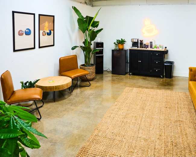 A modern, minimalistic lounge area with a brown sofa, a coffee table, two chairs, potted plants, and a cabinet with a coffee machine against a white wall.
