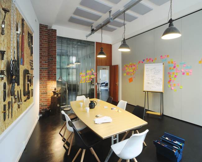 A modern meeting room features a large table surrounded by chairs, with a wall of tools and colorful sticky notes arranged on the adjacent walls.