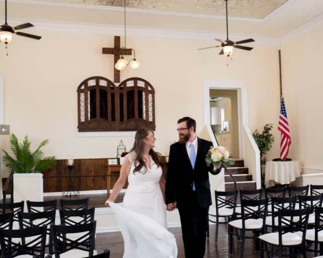 A bride and groom walk hand in hand through a wedding venue with rows of chairs and a cross on the wall.