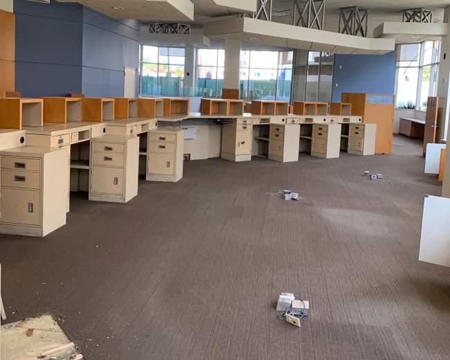 The image shows an empty office space with several desks arranged in rows, some of which have papers and debris scattered on the floor.