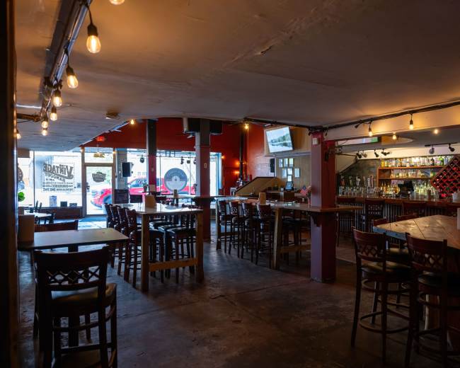 The image shows an empty bar interior with wooden tables and chairs, warm lighting, and a wall lined with shelves of liquor bottles.