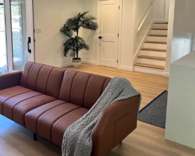 The image shows a modern living area featuring a brown leather sofa, a potted plant, and a staircase leading to an upper level.