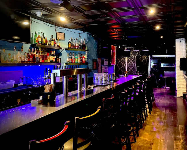 The image shows a modern bar with a long countertop, shelves lined with various liquor bottles, and colorful lighting effects.