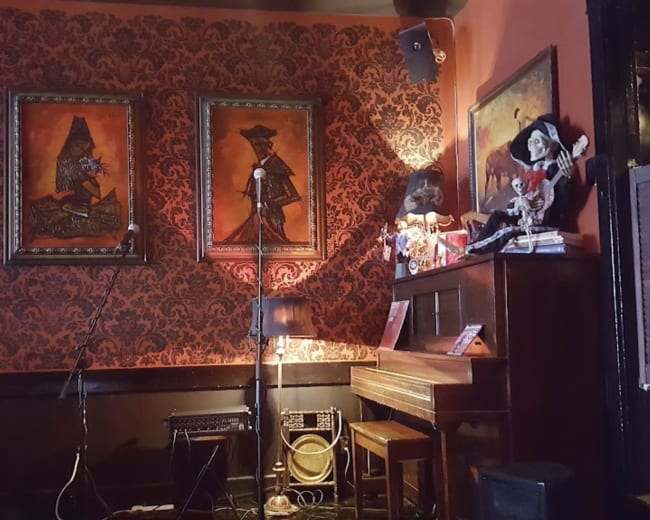 The image shows a corner of a bar with a piano, two framed paintings on the wall, and various decorations, including a microphone and musical equipment.