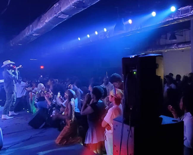 A performer in a hat sings on stage while an audience watches and dances in a dimly lit venue.