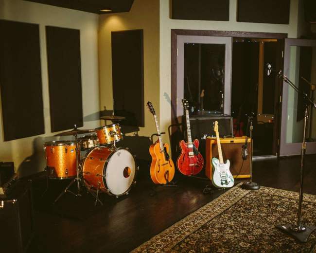 The image shows a music studio setup with a drum kit, several guitars, an amplifier, and a microphone on a stand.