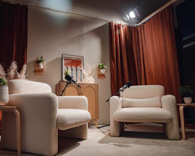The image shows a cozy, minimalist interview setup with two white armchairs, microphones on stands, and decorative plants against a backdrop of orange curtains.