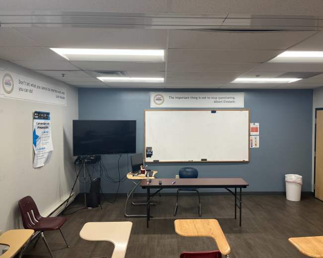 The image shows a classroom with a large monitor, whiteboard, and a central table surrounded by empty chairs.