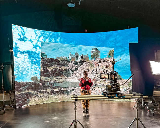 A person stands in front of a large curved screen displaying a landscape, with a camera setup on a tripod nearby in a studio environment.