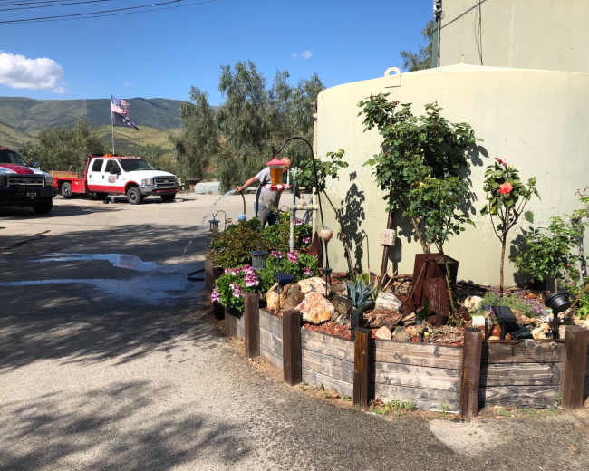 A circular planter filled with various plants and flowers is situated next to a large green water tank, with a red and white truck parked nearby and an American flag in the background.