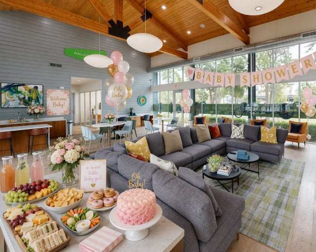 A welcoming indoor space is decorated for a baby shower, featuring a large seating area, a food spread with snacks and sweets, and decorative balloons and banners.