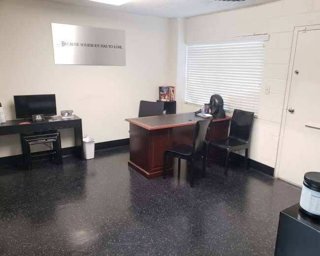 The image shows a small, sparsely furnished office with a wooden desk, two black chairs, a computer setup, and a sign on the wall that reads, "...Because somebody has to lose."