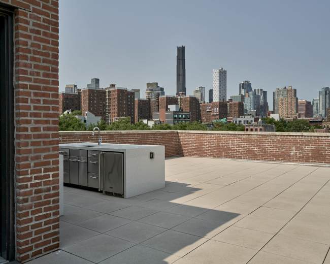 The image shows a rooftop terrace with a modern outdoor kitchen and a skyline view of tall buildings in an urban setting.