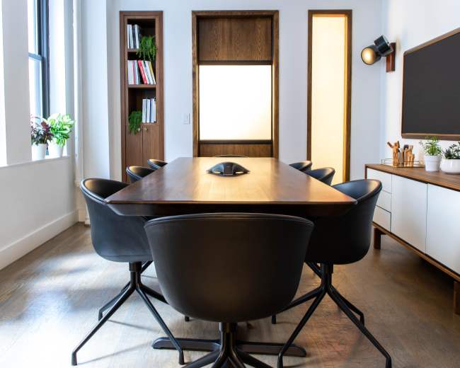 A conference room features a long wooden table surrounded by black swivel chairs, with large windows and minimalist decor.