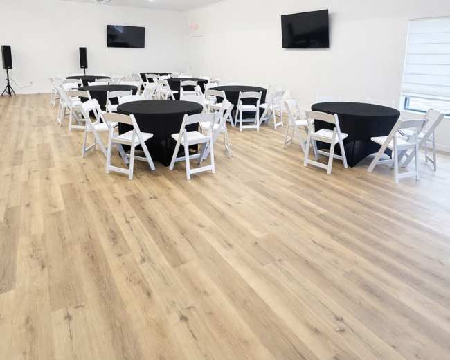 The image shows a spacious event room with several round black tables surrounded by white chairs, wooden flooring, and two wall-mounted televisions.