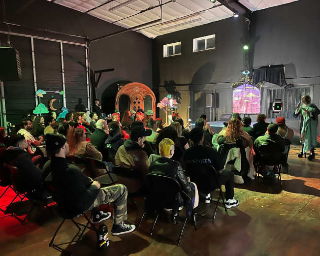 A large audience sits in chairs facing a performer on stage in a dimly lit venue decorated with colorful props.