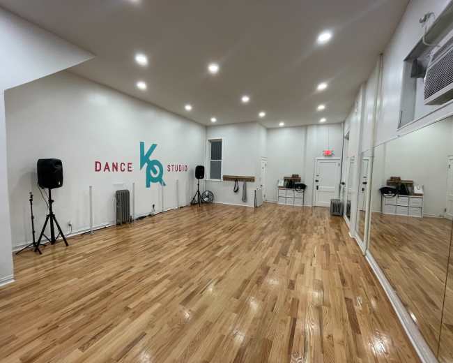 The image shows a spacious dance studio with wooden flooring, mirrors lining one wall, and the words "Dance Studio" in colorful lettering on the opposite wall.