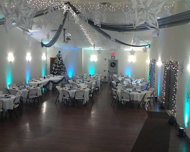 A decorated event space with white tables and chairs arranged in rows, a Christmas tree in the corner, and holiday-themed decorations hanging from the ceiling.