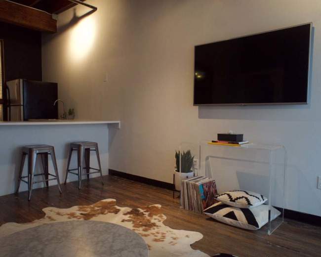 The image shows a modern living space with a kitchen area, a large flat-screen TV mounted on the wall, and a decorative rug on the floor.