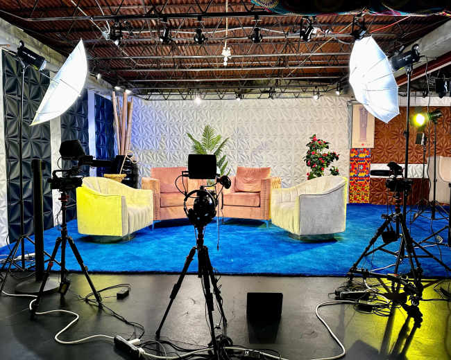 The image shows a television studio setup with two pastel-colored couches on a blue carpet, surrounded by camera equipment and lighting.