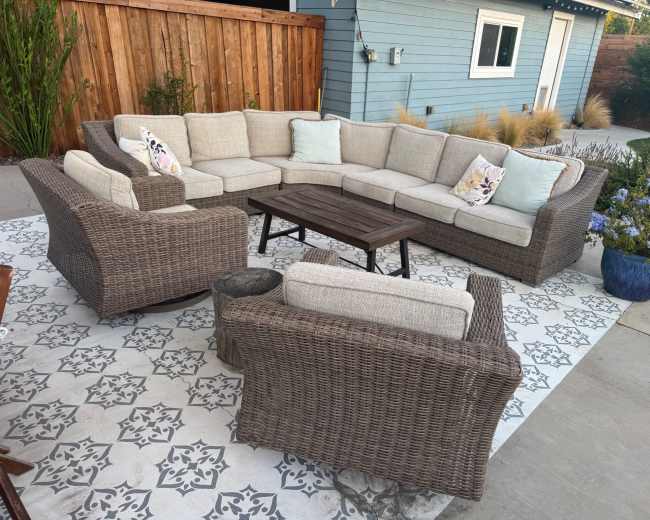 A large, L-shaped outdoor seating arrangement made of wicker is situated on a patterned tile patio, accompanied by smaller wicker chairs and a wooden coffee table.