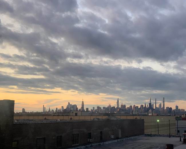 The skyline of a city is visible against a backdrop of clouds and a fading sunset, with various tall buildings, including the Empire State Building, rising in the distance.