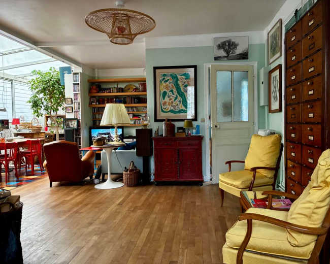 The image shows a cozy living room featuring two yellow armchairs, a wooden floor, a red cabinet, and bookshelves filled with books and decorative items.