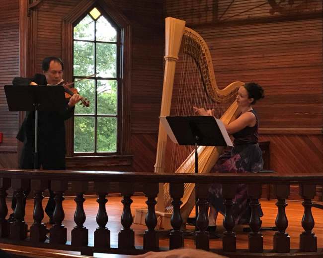 A violinist and a harpist perform together on stage in a wooden-paneled venue.
