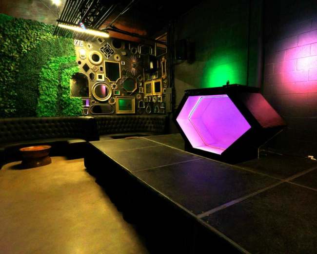 The image shows a small stage with a hexagonal light fixture, surrounded by a seating area and a wall adorned with various framed mirrors and decorations.
