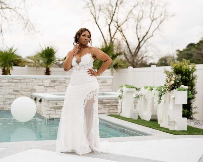 A woman in a white lace wedding dress stands beside a pool with decorative white balloons and a stone wall in the background.