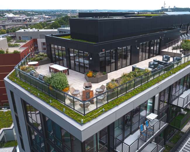 The image shows a modern rooftop terrace featuring a landscaped garden, seating areas, and large glass windows on a multi-story building.