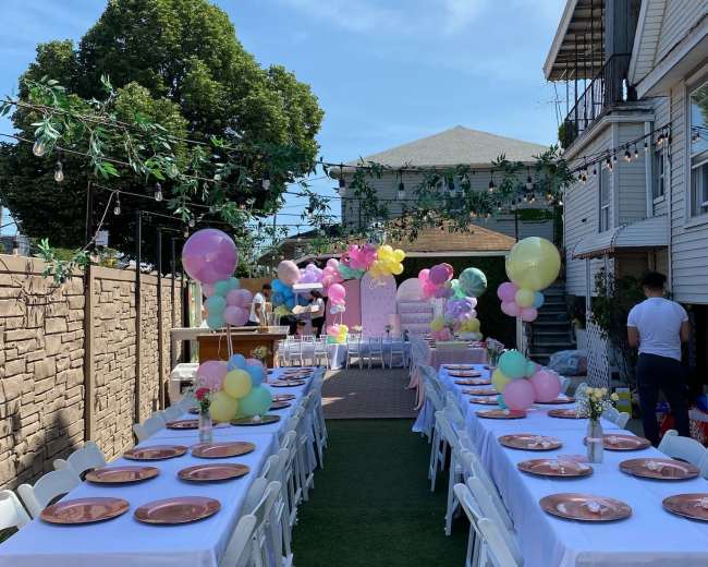 A decorated outdoor space features long tables with white tablecloths set up for a celebration, surrounded by colorful balloons and greenery.