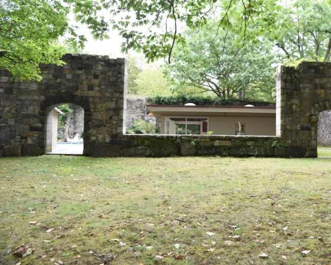 A grassy area framed by two stone wall sections with arched openings, leading to a modern building in the background.