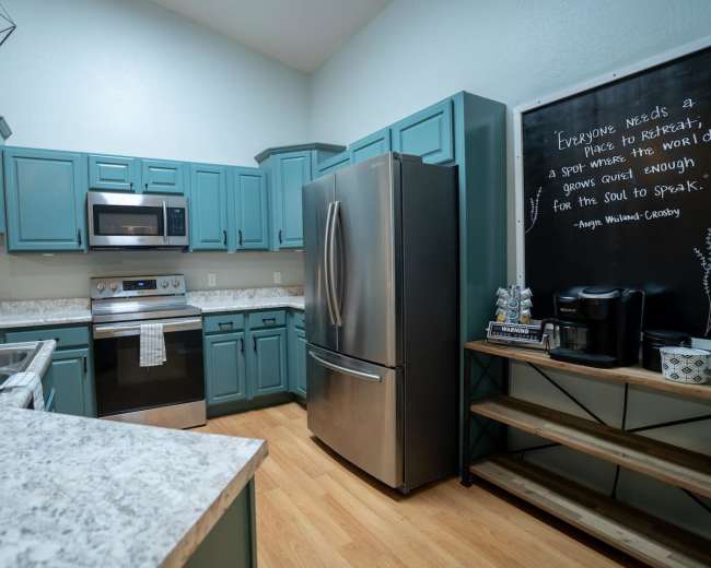 The image shows a modern kitchen featuring teal cabinets, stainless steel appliances, and a chalkboard wall with a quote.