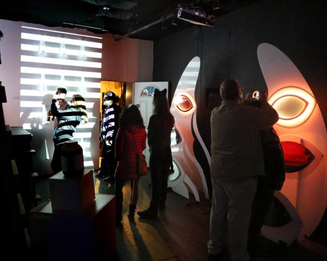 A group of people interacts with an art installation featuring light projections and abstract sculptures in a dimly lit room.