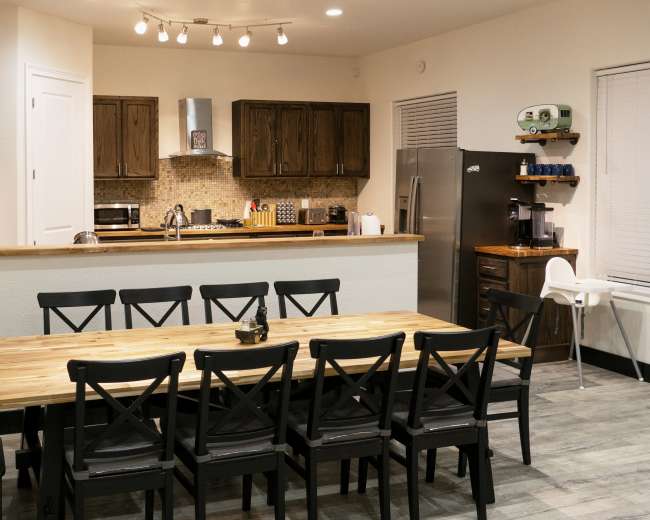 A spacious kitchen and dining area features a large wooden table surrounded by black chairs, with dark wooden cabinetry and modern appliances visible in the background.