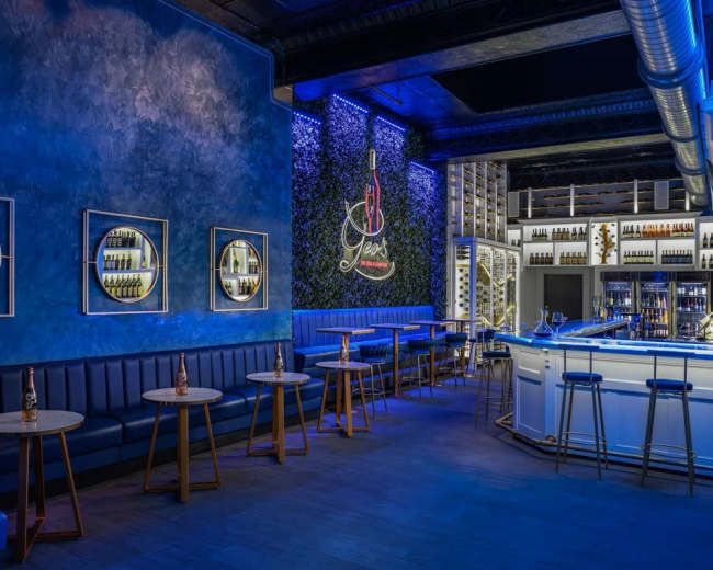 The image shows a modern bar interior with blue walls, circular framed wall art, a white bar counter, and several small tables with chairs.