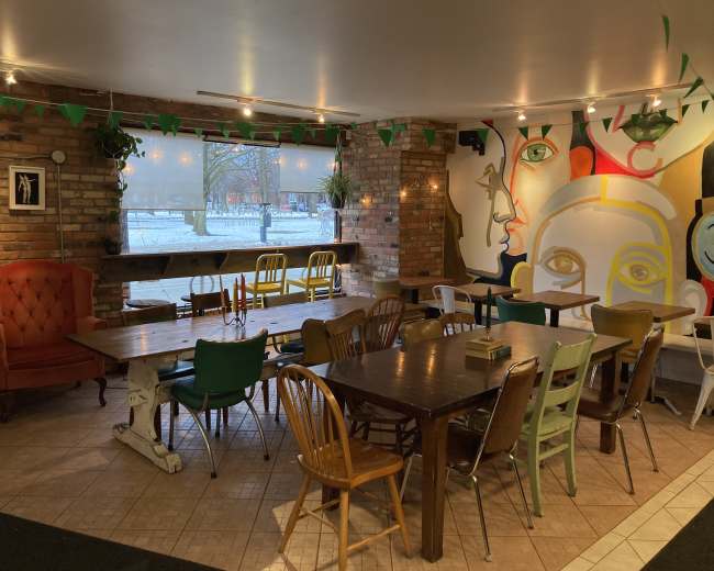 The image shows an interior of a cafe with a mix of wooden and metal tables and chairs, exposed brick walls, and a colorful mural featuring abstract faces.