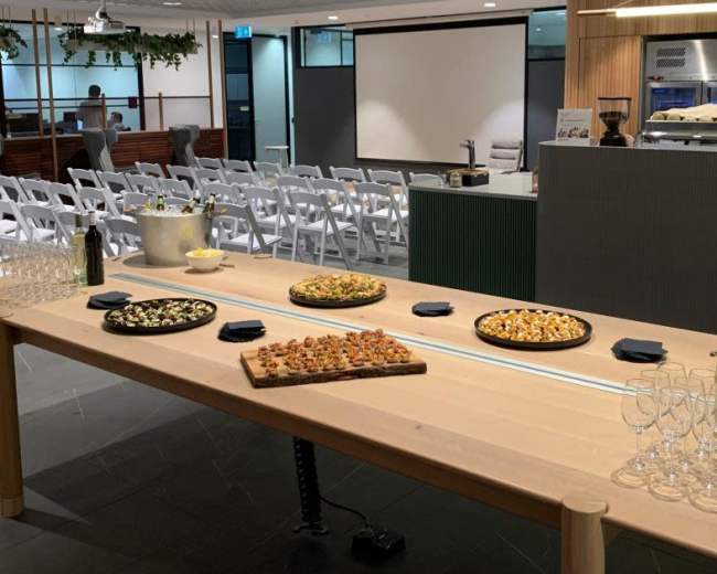 A long wooden table is set with various platters of food and drinks in a spacious room with chairs arranged for an event.