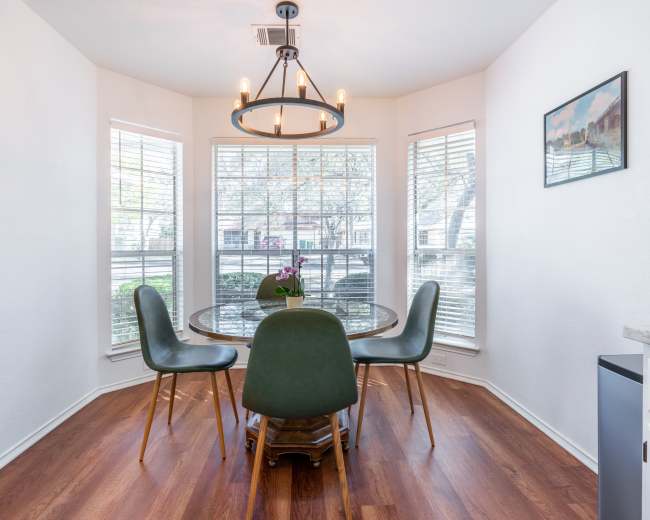 A circular glass dining table with green chairs is set in a sunlit corner surrounded by large windows.