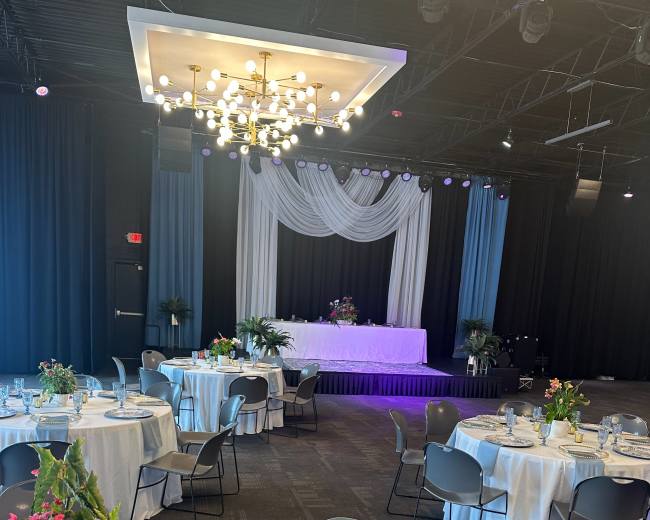 The image shows a banquet hall set up with round tables adorned with white tablecloths, decorative plates, and floral centerpieces, accompanied by a stage draped in white fabric at the front.