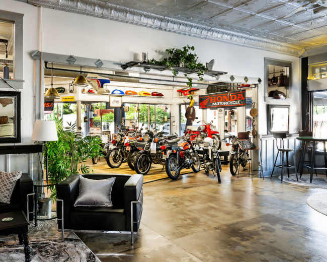 The image shows a stylish interior space with several vintage motorcycles displayed prominently, surrounded by modern furniture and decorative plants.