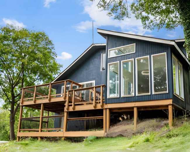 A modern blue house with large windows and a wooden deck is situated on a grassy slope surrounded by trees.
