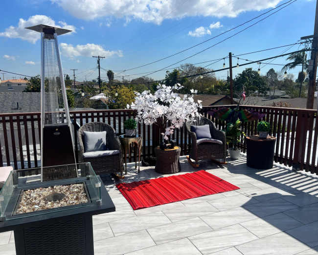 The image shows a rooftop patio with two wicker chairs, a decorative tree, a red rug, and a heater, surrounded by a clear blue sky and power lines.