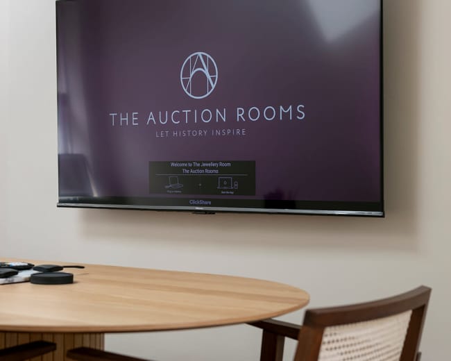 A flat-screen television displaying "The Auction Rooms" logo is mounted on a wall above a round wooden table with wooden chairs.