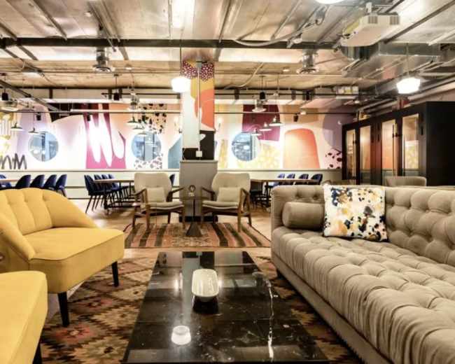 The image shows a modern communal workspace featuring various seating arrangements, colorful wall art, and a combination of tables and chairs.