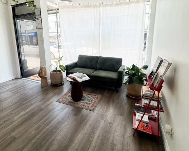 The image shows a cozy interior space with a green sofa, a small wooden table, a bookshelf, and plants near a large window with sheer curtains.