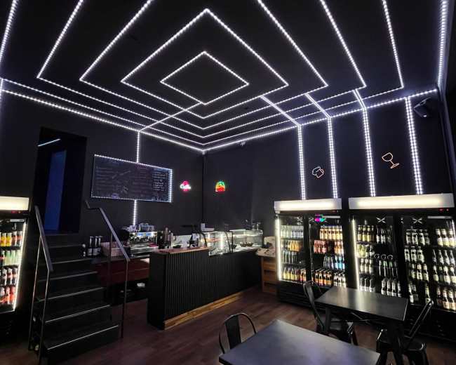 The image shows a modern café with neon lighting, a beverage display, and a staircase leading to another level.