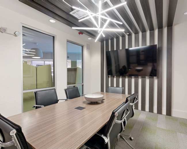 A modern conference room features a large wooden table surrounded by black chairs, with a flat-screen television mounted on a wall beneath contemporary lighting fixtures.