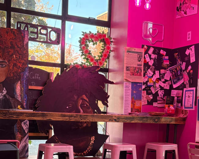 The image shows a vibrant cafe with pink walls, featuring a wooden bar with metal stools and a large painted portrait on the wall.